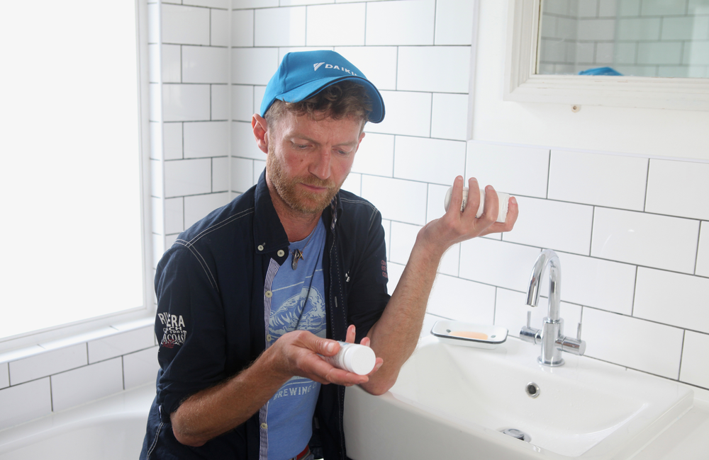 A man in a bathroom holding two bottles of medication and reading one of them.
