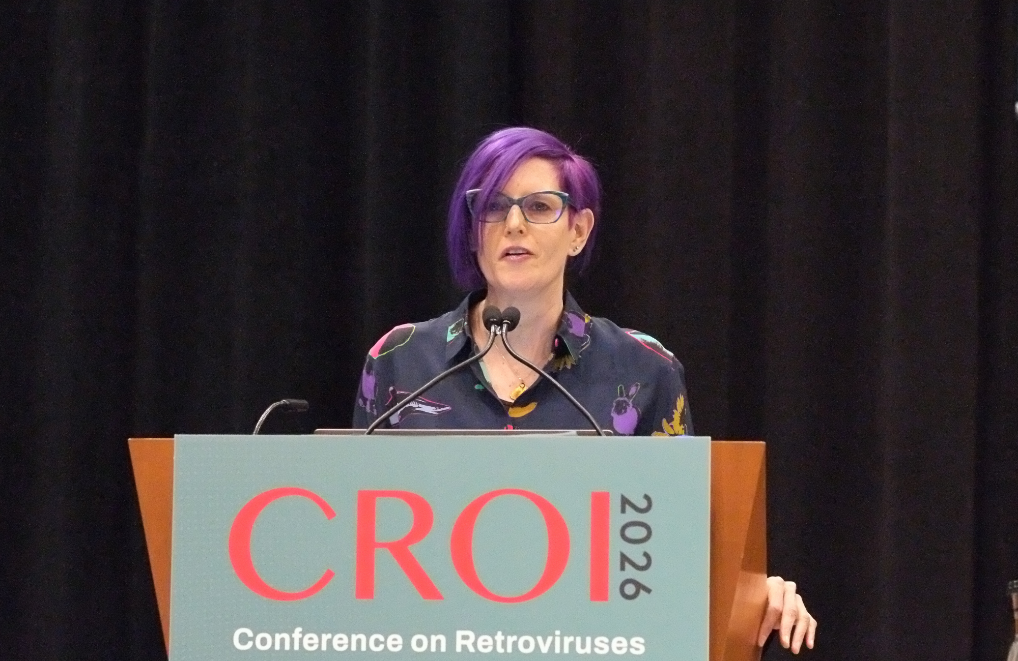 Professor Chloe Orkin at CROI 2026. She is standing behind a lectern talking into a microphone.