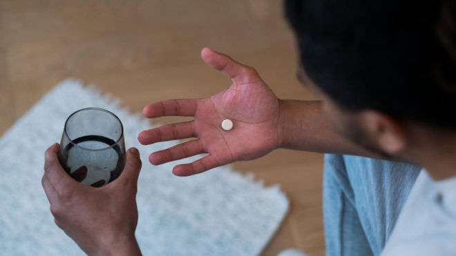 A person holds a small round pill in one hand and a glass of water in their other hand.