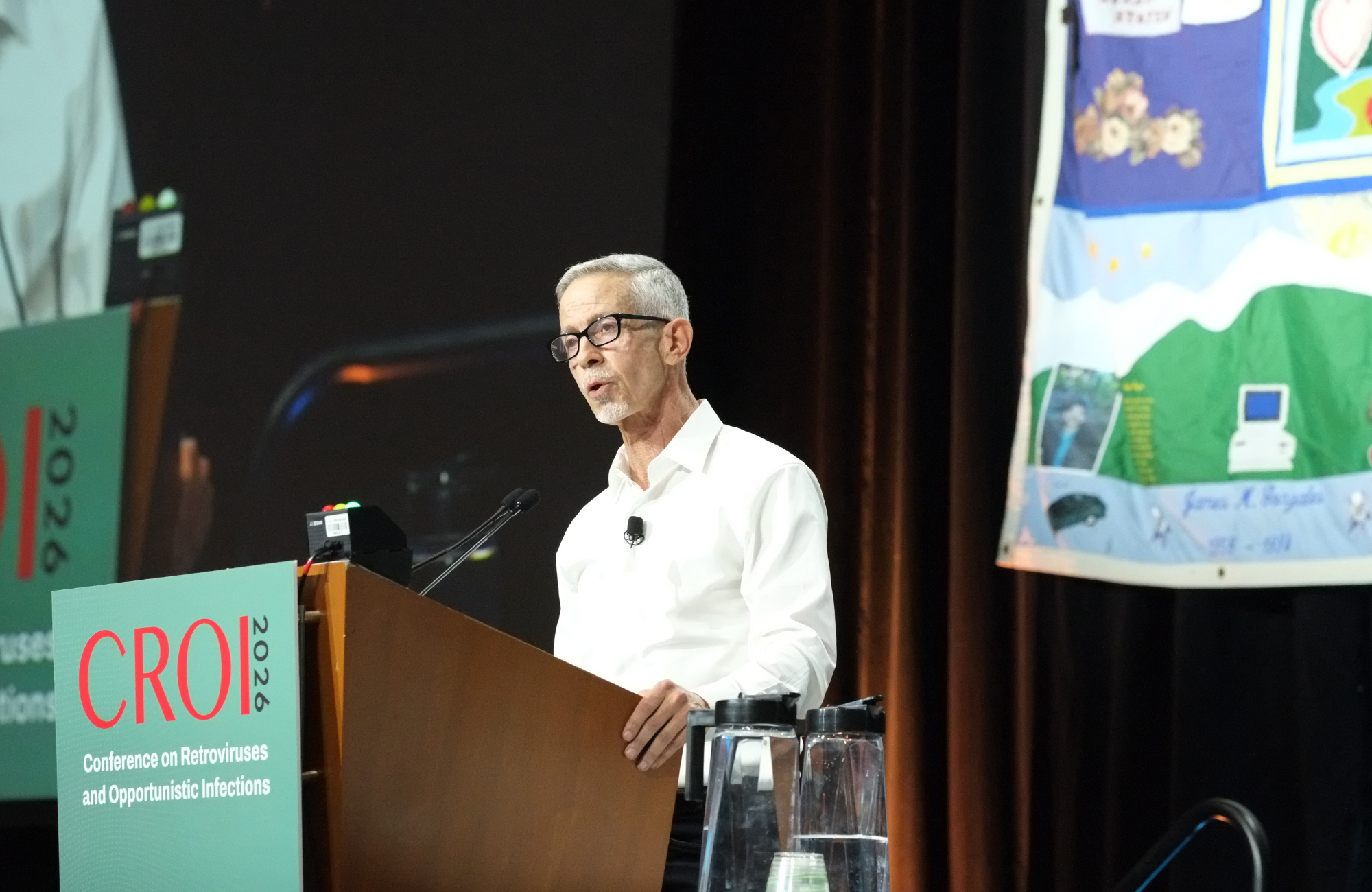 Peter Staley at CROI 2026. He is standing behind a lectern talking into a microphone.
