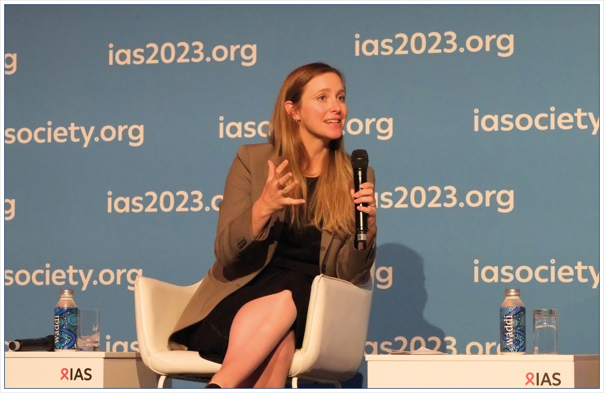 Woman seated on stage talking into a microphone and gesturing.