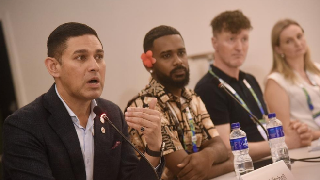 Dr Jason Mitchell at AIDS 2026. He is sitting with three other people (to his left) at a press conference and is talking into a microphone.