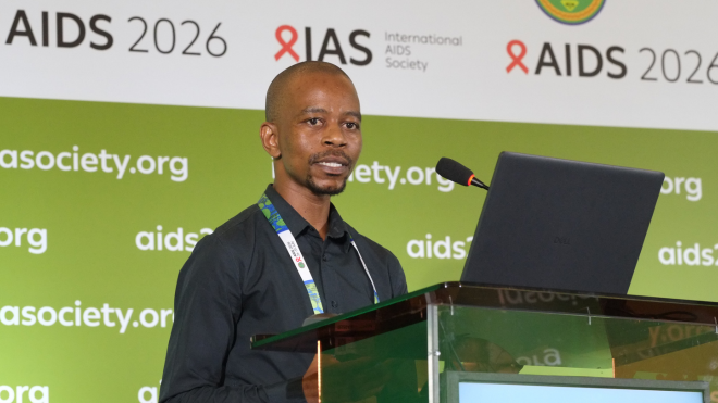 Sanele Mbeje at AIDS 2026. He is standing at a lectern, behind a laptop, talking into a microphone.