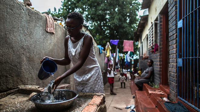 A woman stands outside washing food with a jug of water and a bowl.