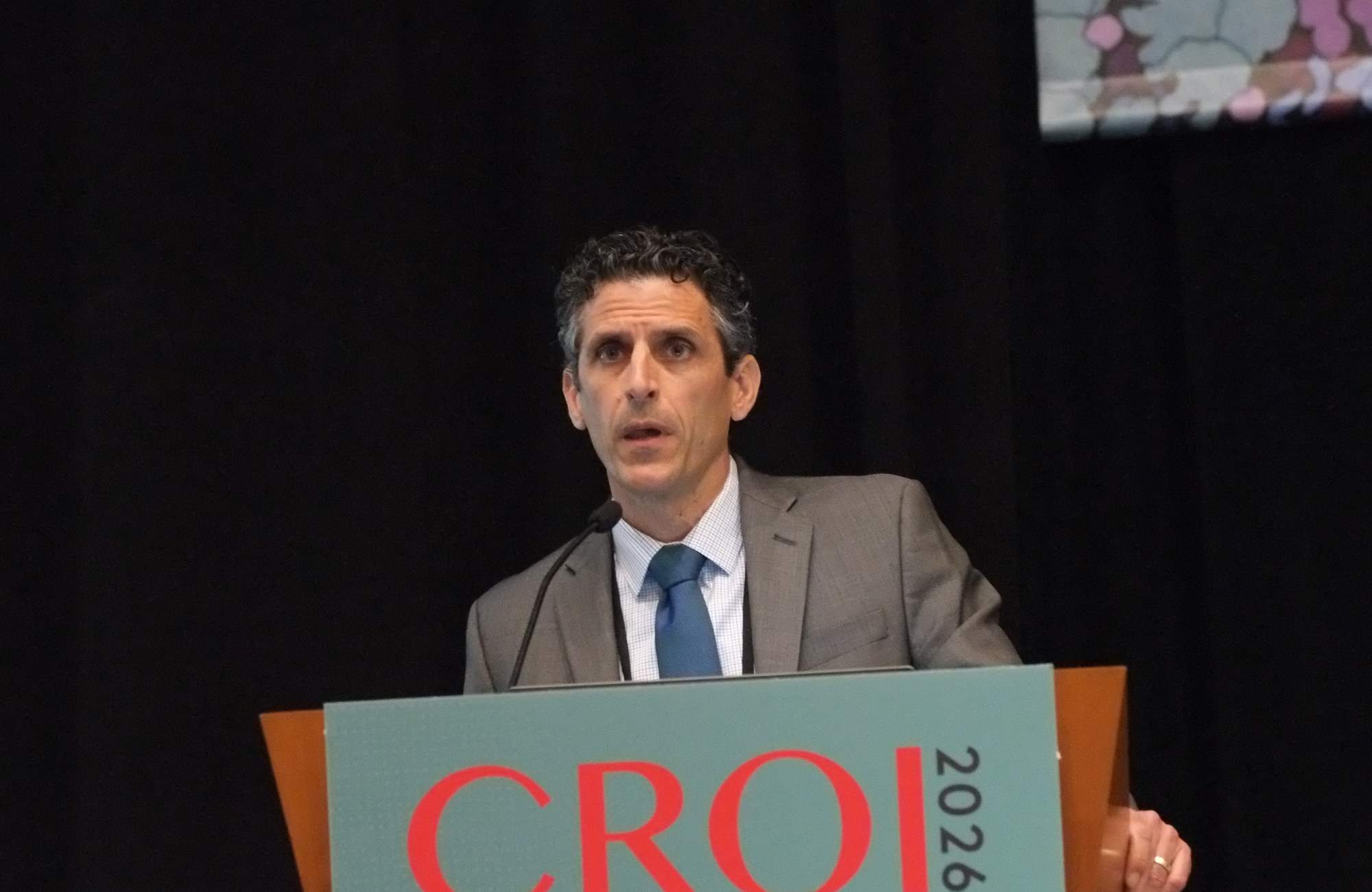 Professor Gabriel Chamie at CROI 2026. He is standing behind a lectern, talking into a microphone.