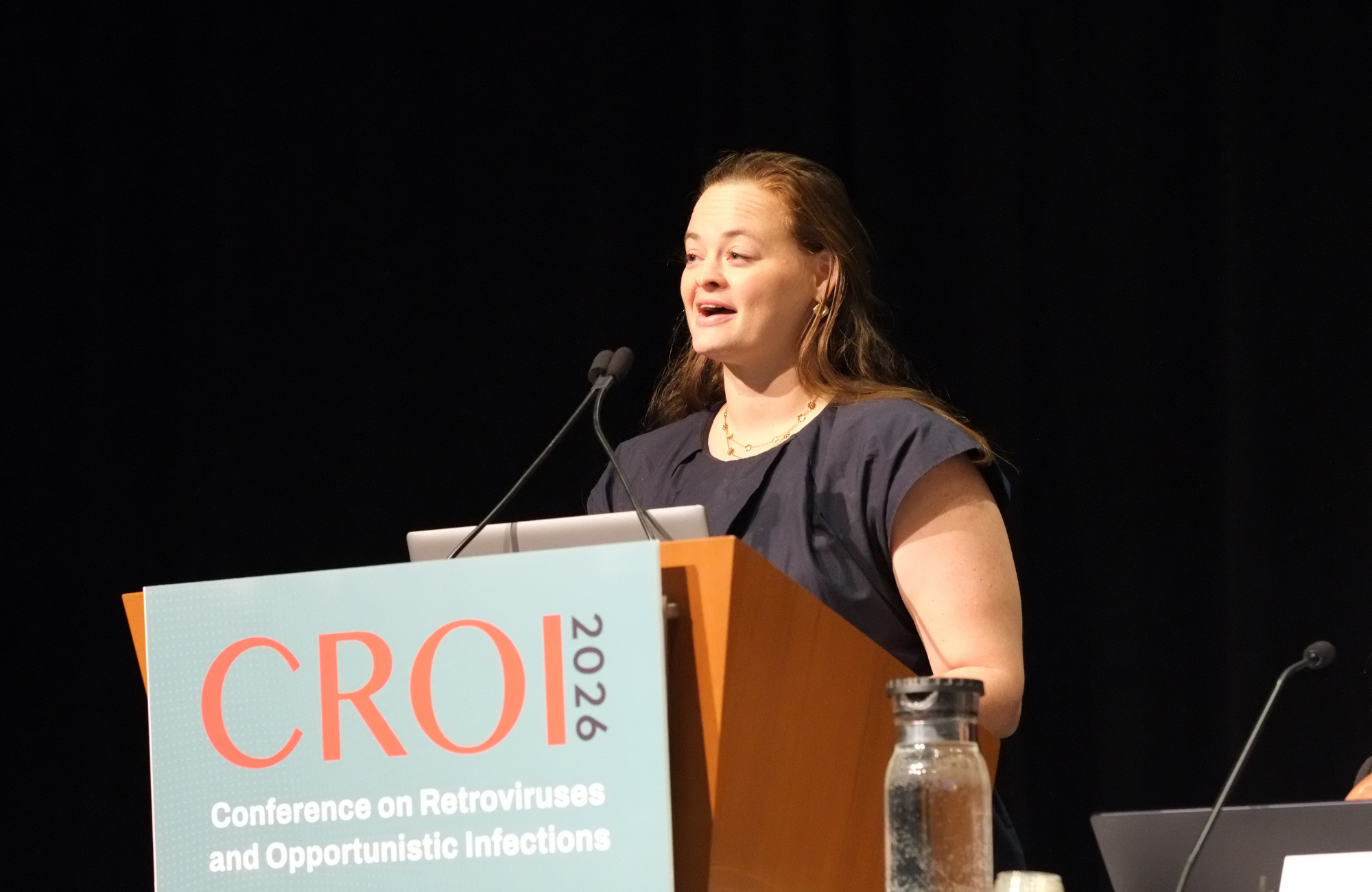 Dr Gabriela Cromhout at CROI 2026. She is standing behind a lectern, talking into a microphone.