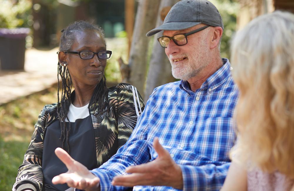 A picture of three people in conversation outside on a sunny day. All three people are sat down. On the left is a Black woman who looks interested in the person next to her. This is an older white man wearing glasses and a baseball cap. He is saying something that is making him smile and using his hands to express himself. In the foreground we can see the back of somebody's head who is facing the man. This person has long, blonde, wavy hair. he image has a pink and purple filter over it. 