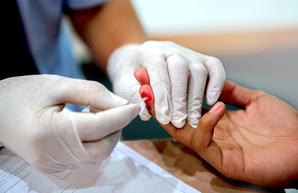 A person wearing a surgical glove holds the hand of somebody else. This person is having their finger pricked with a fine needle. The needle has drawn a small amount of blood which is visible. The image has a pink and purple filter over it. 