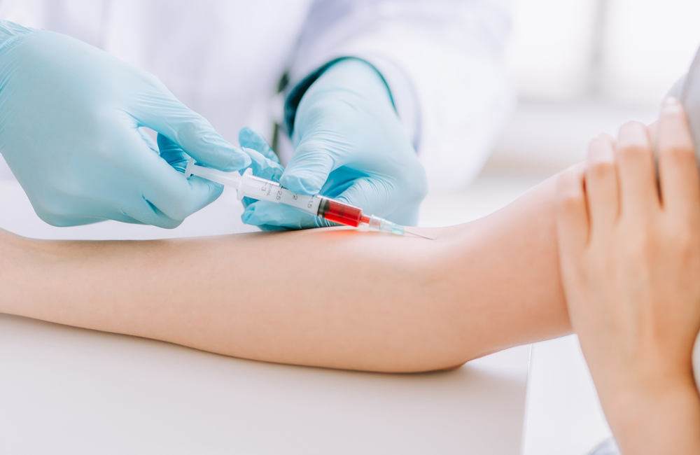 A close up of a person's arm while they are having their blood taken. The person taking the blood is wearing surgical gloves. We can see some of the blood is already in the tube. 