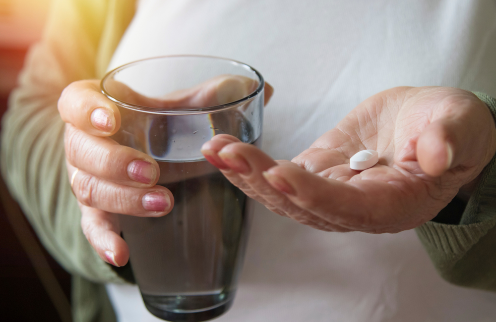 Image: carballo/Shutterstock.com A close up on a person's hands. One hand is holding some medication, the other hand is holding a small glass of water. The image has a pink and purple filter over it.