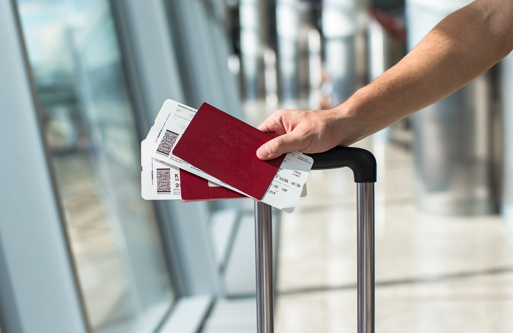 A person holding onto a suitcase handle along with two red passports and plane tickets, at an airport.