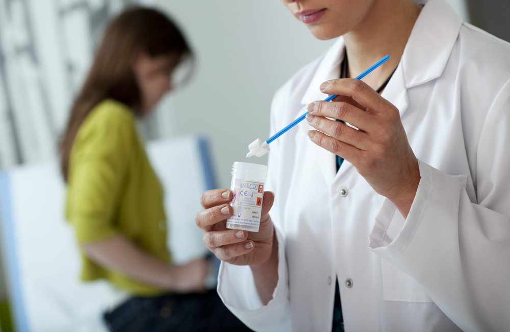 In the foreground, a healthcare professional in a white coat is putting a swab into a container. In the background, blurred, we can see a woman with long hair sat on a clinic bed. 