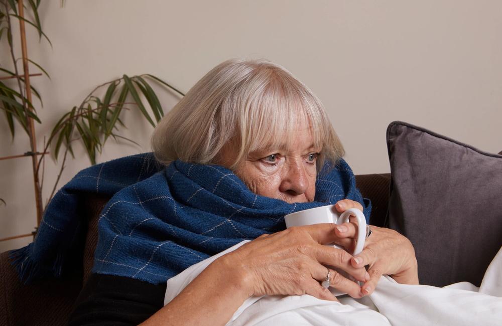 Image credit: Domizia Salusest | www.domiziasalusest.com An older woman sitting on the sofa with a blanket around her shoulders, looking tired. She is holding a mug.