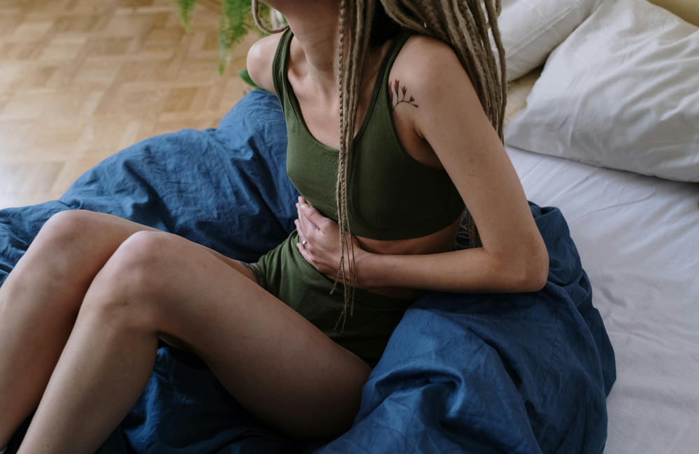 cottonbro studio/Pexels.com. Image is for illustrative purposes only. A woman sits on a bed on a blue blanket holding her stomach.