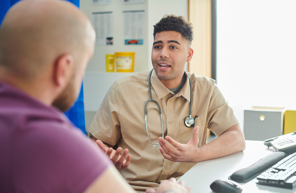A doctor wearing a light brown shirt and a stethoscope around his neck sitting at a desk and talking to a patient who's wearing a purple t-shirt.