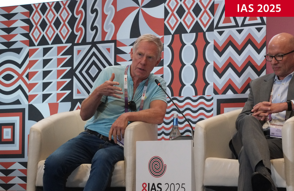 Dr Graeme Moyle at IAS 2025. He is sitting on a grey chair and talking into a microphone. He's wearing a pale blue top. 