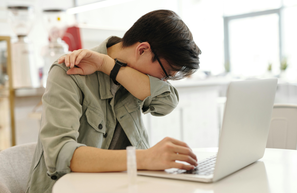 A man sits at a desk in front of a laptop and is coughing into his arm.