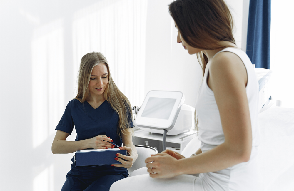 A woman sits in a doctor's office on a bed and talks to a doctor.