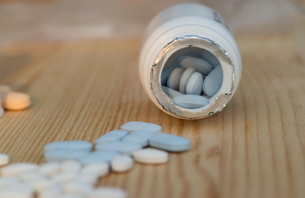 A white pill bottle is tipped over with blue pills spilling out on to a wooden table., along with white pills.