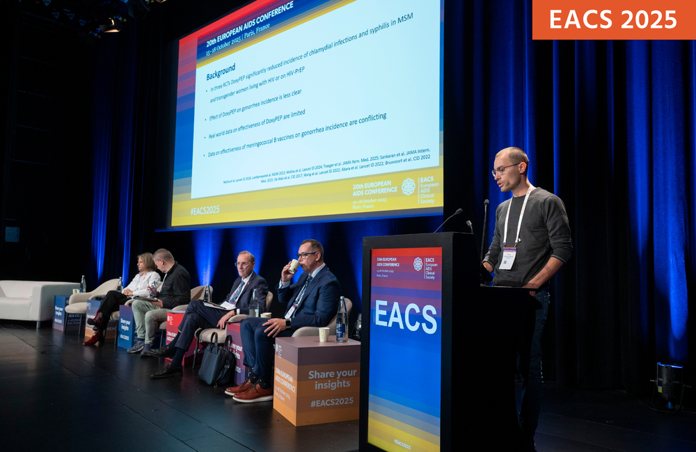 Dr David Wimmersberger presenting at EACS 2025. He is talking into a microphone at a podium with a screen and panel of speakers to his left.