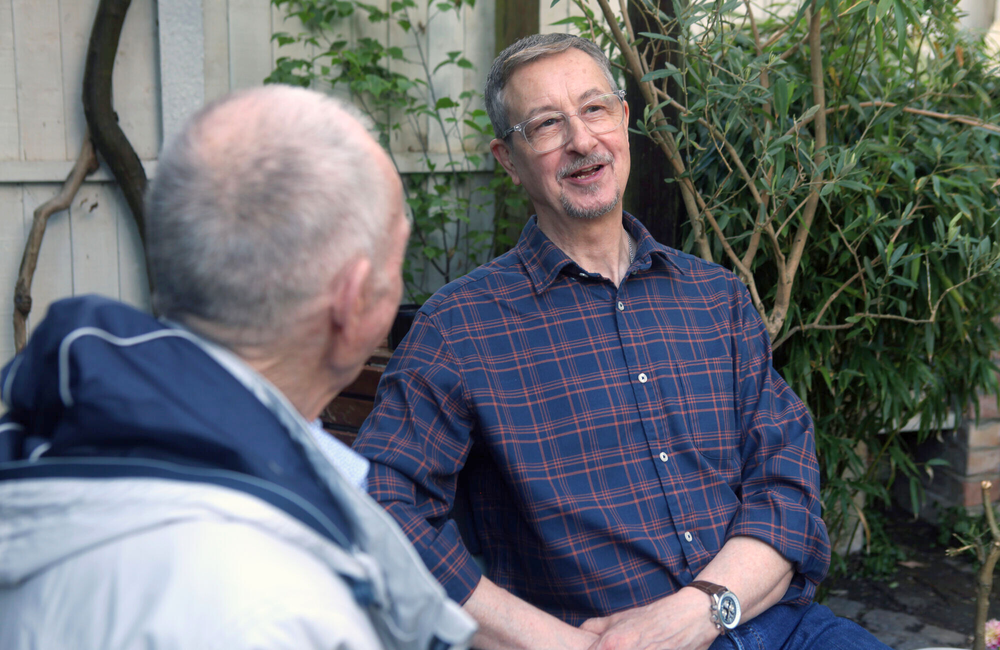 Two older men talk to each other in a garden.