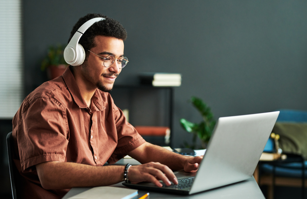 A man sits at a desk using a laptop and wearing white headphones.
