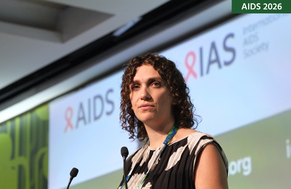 Elise Lankiewicz at AIDS 2026. Elise is standing behind a microphone, with a screen behind her.