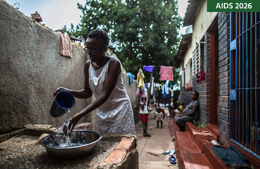 A woman stands outside washing food with a jug of water and a bowl.