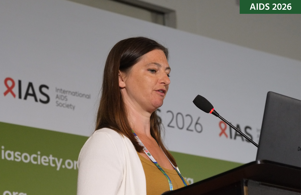 Florence Bascombe at AIDS 2026. She is standing at a lectern, talking into a microphone.