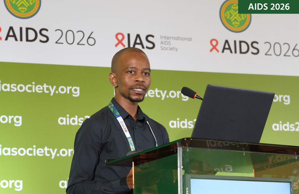 Sanele Mbeje at AIDS 2026. He is standing at a lectern, behind a laptop, talking into a microphone.